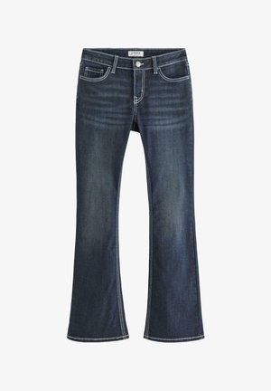 Ung person stående med hånden i lommen iført hvid kortærmet skjorte, mørkeblå jeans og hvide sneakers mod en ensfarvet baggrund.
