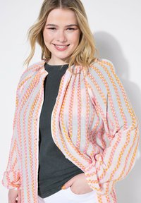 Lehká, průsvitná, košile na knoflíky s růžovými, oranžovými a šedými pruhy; má dlouhé rukávy a uvolněný střih přes tmavý top.