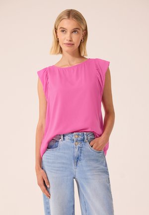 Kvinde med blondt hår iført en knaldpink ærmeløs bluse, der er stoppet ned i lyseblå jeans med høj talje, med én hånd i lommen.