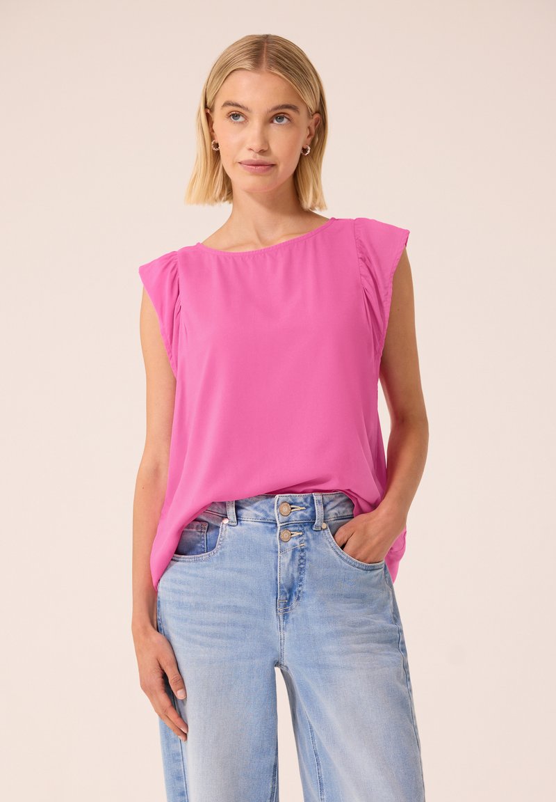 Femme aux cheveux blonds portant un chemisier sans manches rose vif, rentré dans un jean taille haute bleu clair, une main dans la poche.