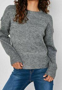 Femme portant un pull en tricot gris à manches longues avec des trous pour les pouces et un jean bleu, debout les mains près des hanches devant un fond uni.