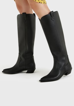 Černé kožené kozačky sahající po kolena s nízkými podpatky a špičatou špičkou, nošené s krátkou žlutou sukní.