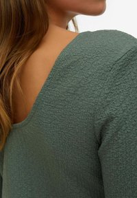 Groene gestructureerde top met een brede halslijn, voorzien van een gekreukt materiaal en een lichte glans, met een close-up van de achterkant en schoudergebied.