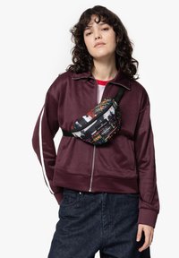 Černý ledvinkový batoh s barevnými graffiti vzory; nošený diagonálně přes vínově červenou zipovou bundu a tmavé džínové kalhoty. Texturovaný povrch látky.