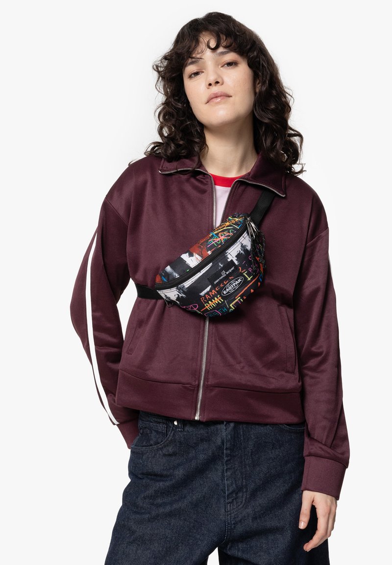 Marsupio nero con motivi grafici colorati; indossato in diagonale su una giacca a zip borgogna e pantaloni in denim scuro. Finitura in tessuto testurizzato.