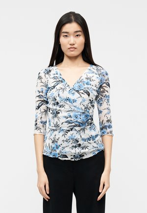 Γυναίκα με μακριά σκούρα μαλλιά που φοράει λευκή, μπλε και μαύρη μπλούζα τύπου wrap με λουλούδια και μανίκια τριών τετάρτων, καθώς και μαύρο παντελόνι, στέκεται μπροστά σε λευκό φόντο.