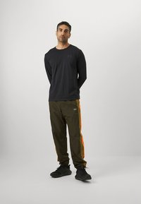 Svart långärmad tröja kombinerad med olivgröna byxor med starkt orange sidostripar. Tyget ser slätt och lätt ut.