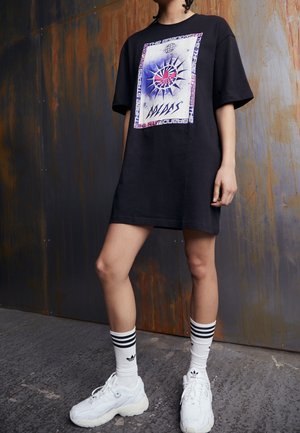 Osoba nosząca oversize czarną sukienkę w stylu koszulki z grafiką, białe skarpetki Adidas z czarnymi paskami i białe masywne trampki stojąca w pomieszczeniu.