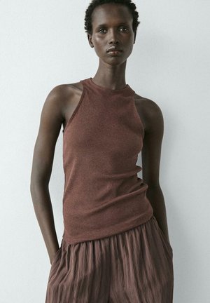 Femme aux cheveux courts et bouclés portant un haut marron sans manches et un pantalon marron ample, debout devant un mur clair uni.