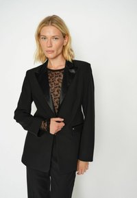 Schwarzer maßgeschneiderter Blazer mit satinierter Revers, taillierter Form und Ein-Knopf-Verschluss. Trägt man über einer transparenten, gemusterten Bluse.
