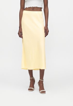 Γυναίκα που φοράει μια μίντι φούστα σε ανοιχτό κίτρινο χρώμα, άσπρο κοντό τοπ και μπεζ πέδιλα με λουράκι στον αστράγαλο, στέκεται μπροστά σε ένα απλό λευκό φόντο.