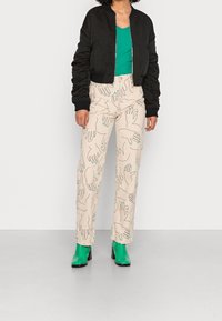 Černá bomber bunda, zelený top s výstřihem do V, krémové kalhoty s modrými otisky rukou. Zelené kotníčkové boty na blokovém podpatku doplňují vzhled.