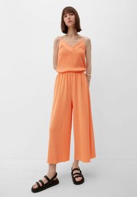 Oranje jumpsuit met een V-hals en dunne spaghettistrings, met wijde broekspijpen en een cinched tailleband. Zwarte sandalen met meerdere banden.