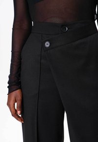 Zwarte high-waisted broeken met een gevouwen voorkant, voorzien van twee zwarte knopen en een strakke, gestructureerde uitstraling. De stof lijkt glad en goed gesneden.