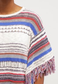 Top a righe in maglia bianco, blu, rosso e grigio. Presenta dettagli a lavorazione aperta e maniche con frange per aggiungere texture ed elementi di design intricati.