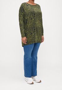 Långärmad tunika i olivgrön med svart leopardmönster, parat med blå jeans och vita sneakers. Avslappnad passform och mjuk textur.