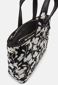 Czarna i biała torba na ramię z kwiecistym wzorem, wykonana z tkaniny, z gładkimi, czarnymi skórzanymi uchwytami, złotym zamkiem błyskawicznym i przestronnymi przegródkami wewnętrznymi.