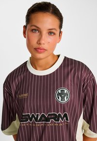 Mørkelilla trøje med hvide lodrette striber, kortærmet og rund hals. Har et logo og mærketekst i kontrastfarver.