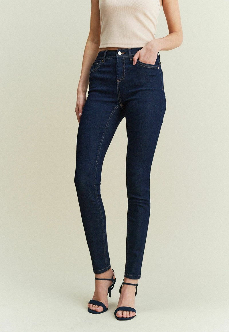 Tmavě modré skinny džíny s hladkou texturou, s designem pěti kapes, vpředu se zipem a zapínáním na knoflík. Stylizováno s černými sandály na podpatku.