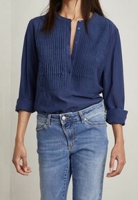 Blusa blu navy con pieghe verticali sul davanti, scollo rotondo e chiusura con bottoni, abbinata a jeans azzurri chiari.