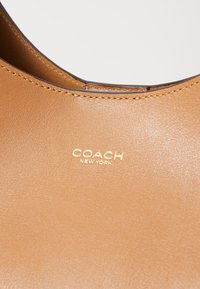 Lichte leren handtas met een gladde textuur, voorzien van een gouden "COACH NEW YORK"-logo en contrasterende stiksels langs de randen.
