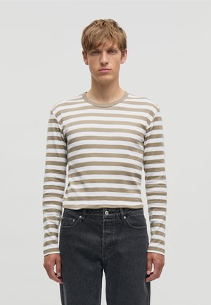 Joven con cabello castaño claro, vestido con una camisa de manga larga a rayas beige y blanca y jeans negros, de pie contra un fondo claro y liso.