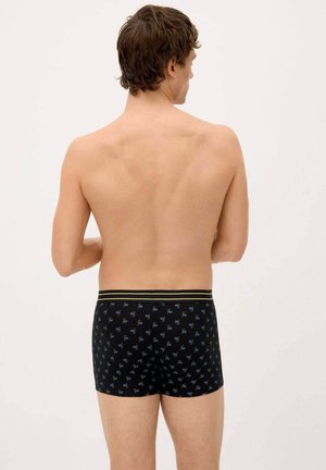 Hombre visto desde atrás, vistiendo bóxer negros con pequeños patrones azules y una cintura con franjas doradas, sobre un fondo liso.