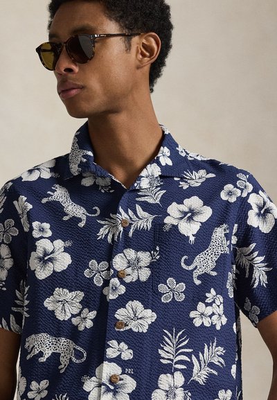 Granatowa koszula z krótkim rękawem, ozdobiona białymi wzorami kwiatowymi i panterkowymi, wykonana z teksturowanej tkaniny, z brązowymi guzikami na klasycznym kołnierzyku.
