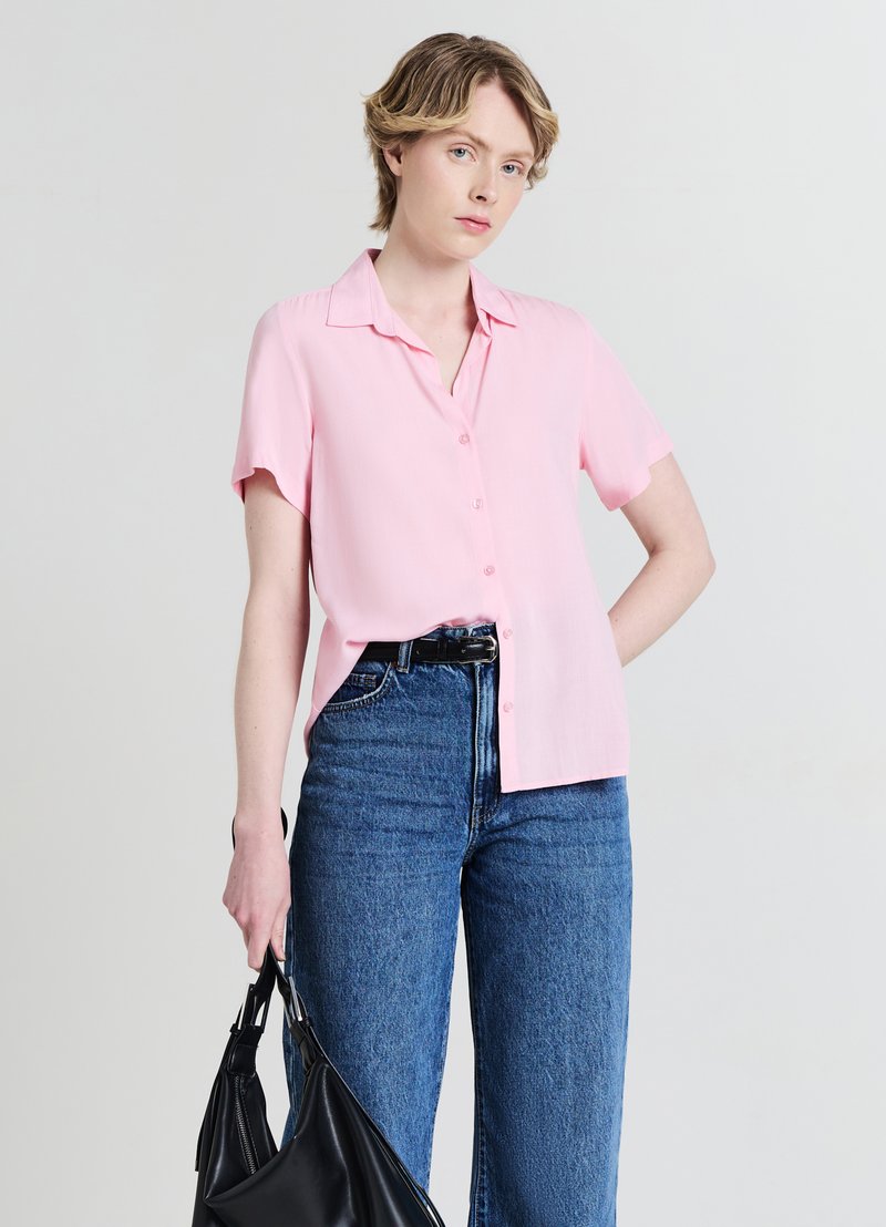 Chemise rose clair à manches courtes avec des boutons et une texture subtile, associée à un jean bleu taille haute et un sac à main noir.