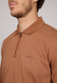 Polo shirt marron avec un col zippé, fabriqué dans un tissu doux. Présente un logo discret sur le côté gauche. Texture lisse, design ajusté.