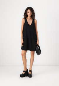 Robe noire sans manches avec un décolleté en V, deux poches avant et une coupe décontractée. Le mannequin tient un sac noir et porte des chaussures à plateforme noires et épaisses.