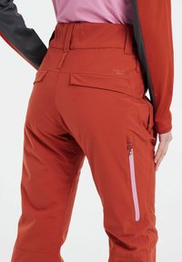 Pantaloni da sci rossi con vita alta, tasca laterale con zip in rosa e una texture liscia. Presenta un logo sopra la tasca posteriore.