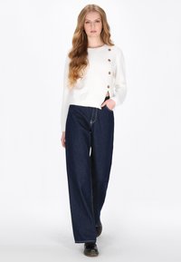 Hvid, blød, cropped sweater med tre brune knapper, parret med brede, mørkeblå denim jeans og sorte sko. Simpelt, afslappet outfit.