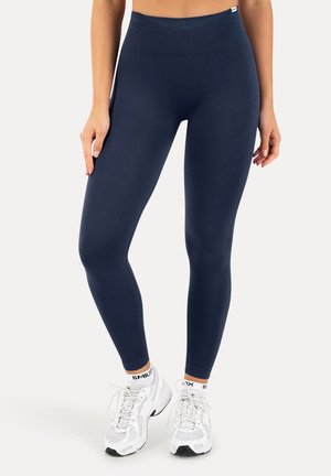 Kvinde iført mørkeblå leggings med høj talje og hvide sneakers med hvide sokker, stående mod en ensfarvet lys baggrund.