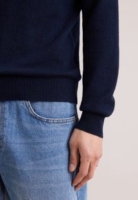 Pull en laine bleu marine avec des poignets côtelés, associé à un jean en denim bleu clair. Le pull présente une texture douce et une coupe ajustée.