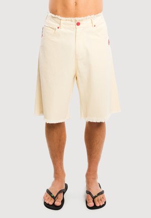 Mandlig model iført off-white knælange denimshorts med frynser og sorte flip-flops, stående mod en ensfarvet baggrund.