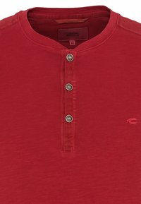 Maglietta henley rossa a maniche corte in cotone, con una chiusura a bottoni composta da tre bottoni argentati e un piccolo logo sul lato.
