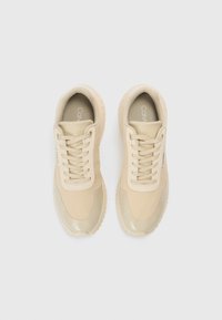 Beżowe buty sportowe z gładką cholewką, siateczkowym materiałem, okrągłym noskiem i teksturowaną gumową podeszwą. Posiadają sznurowany fason i markowe detale.