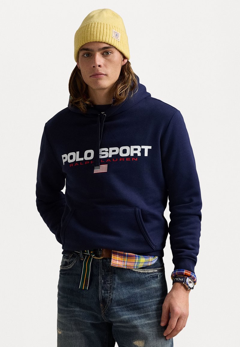 Sudadera de marina con el logo "POLO SPORT" en blanco y rojo, que cuenta con un bolsillo frontal. Combinada con vaqueros desgastados y una camisa de cuadros multicolores.