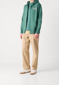 Felpa verde con cappuccio e zip, dotata di tasche frontali, indossata sopra una maglietta coordinata. Abbinata a pantaloni beige e scarpe da ginnastica bianche con suola marrone.