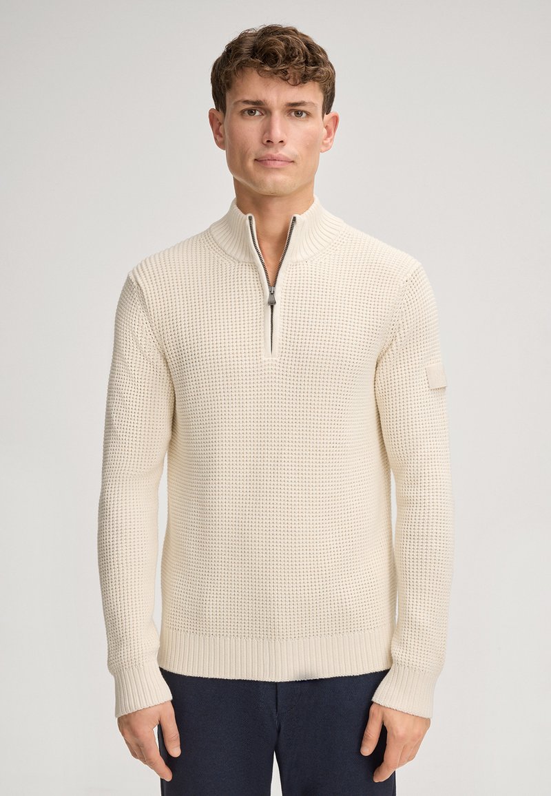 Pullover in maglia beige chiaro con trama, collo con zip e polsini a coste. Presenta un piccolo emblema sulla manica sinistra. Tessuto morbido, vestibilità regolare.