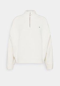 Weißer gestrickter Pullover mit hohem Kragen, halbem Reißverschluss vorne und gerippten Bündchen. Bewusst hat er ein kleines Logo auf der linken Brustseite.