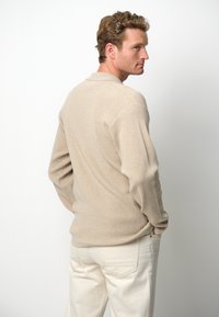 Beige Strickpullover mit Rippstruktur, langen Ärmeln und kragenlosem Ausschnitt, kombiniert mit hellfarbigen Hosen.
