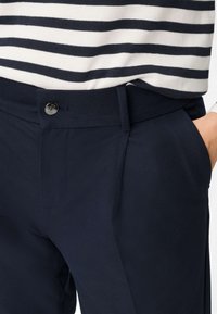 Marineblauwe, op maat gemaakte broek met een gestructureerde stof, voorzien van een knoopsluiting aan de voorkant en zijzakken, gecombineerd met een wit gestreept overhemd.