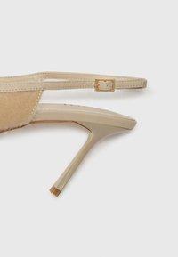 Beige augstpapēžu sandales ar zamša paneļiem, plānu potītes siksniņu ar zelta sprādzi un uzpuntētu pirkstu dizainu. Gluda tekstūra un eleganta forma.