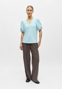 Haut bleu clair à manches courtes et col en V avec manches bouffantes, associé à un pantalon brun ample et des chaussures noires. Tissu lisse et design simple.