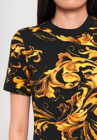 Zwart T-shirt met een gedurfd geel bloemen- en barokpatroon, ronde halslijn en korte mouwen. Merk tekst verschijnt op de stof.