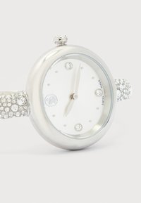 Στρογγυλό ασημένιο ρολόι χειρός με λευκό καντράν, ενδείξεις ώρας από κρύσταλλο στις θέσεις 12, 3, 6 και 9, και λουράκι με διακοσμητικά κρύσταλλα, δείχνει 12:05.