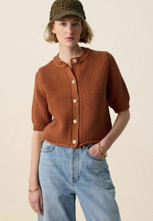 Femme portant un cardigan court en maille marron à manches courtes avec des boutons dorés, un jean bleu clair, une casquette imprimé léopard et des bijoux dorés.