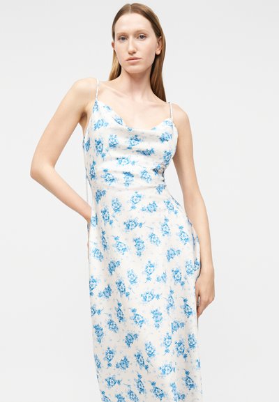 Γυναίκα στέκεται με το χέρι στη μέση, φορώντας ένα λευκό αμάνικο φόρεμα με λεπτές τιράντες και μπλε λουλουδάτο σχέδιο πάνω σε απλό φόντο.